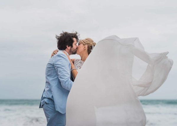 Bride and groom kissing on a beach, wedding dress being blown by the wind by John Barwood Photography